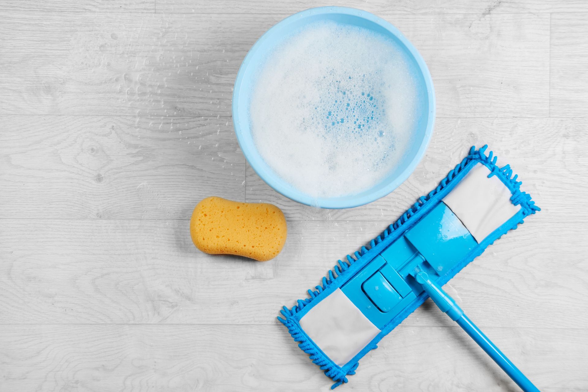Blue mop, soapy water, and sponge on light-colored wood floor, ready for cleaning.