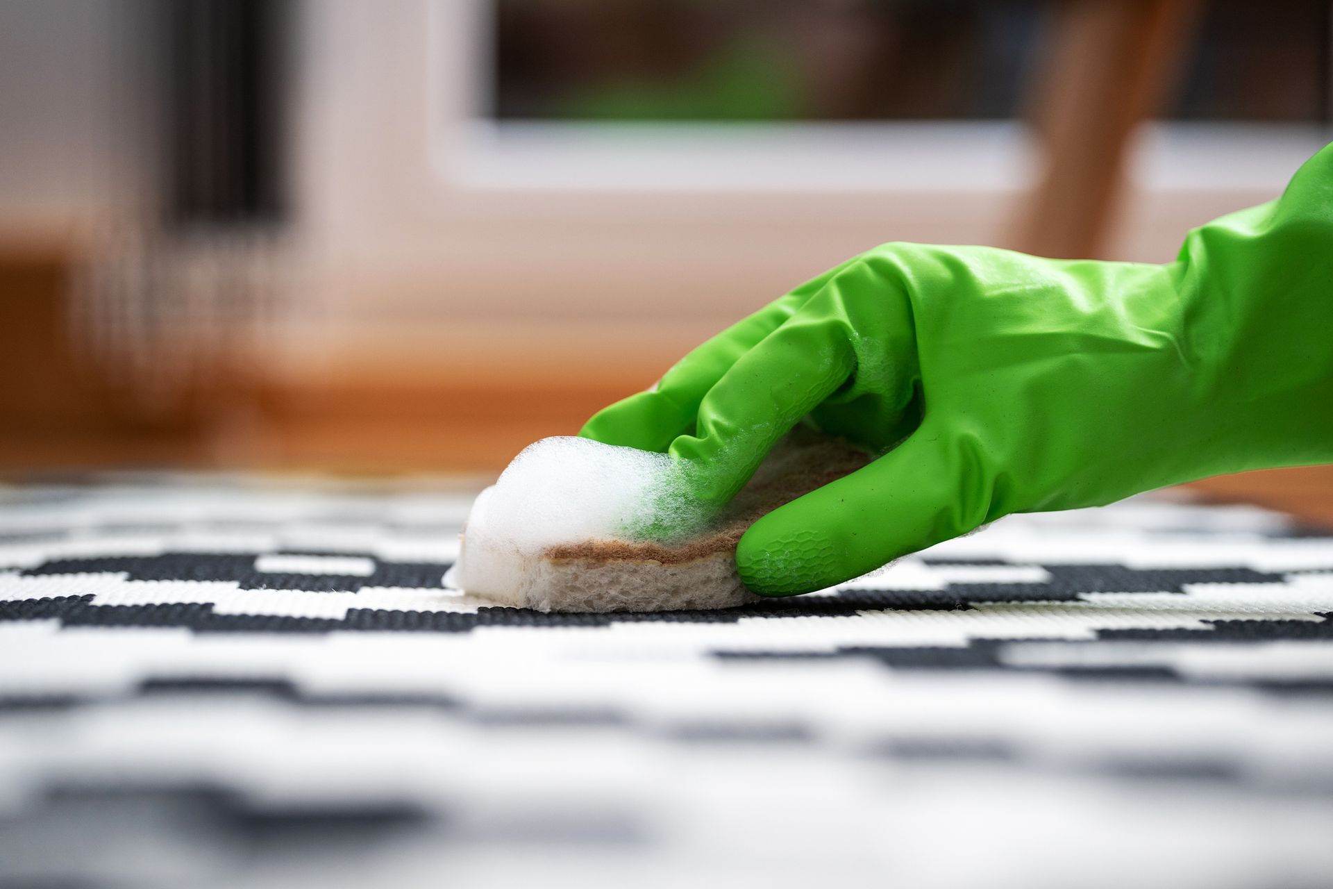 Hand in green glove cleaning a black and white patterned carpet with a soapy sponge.