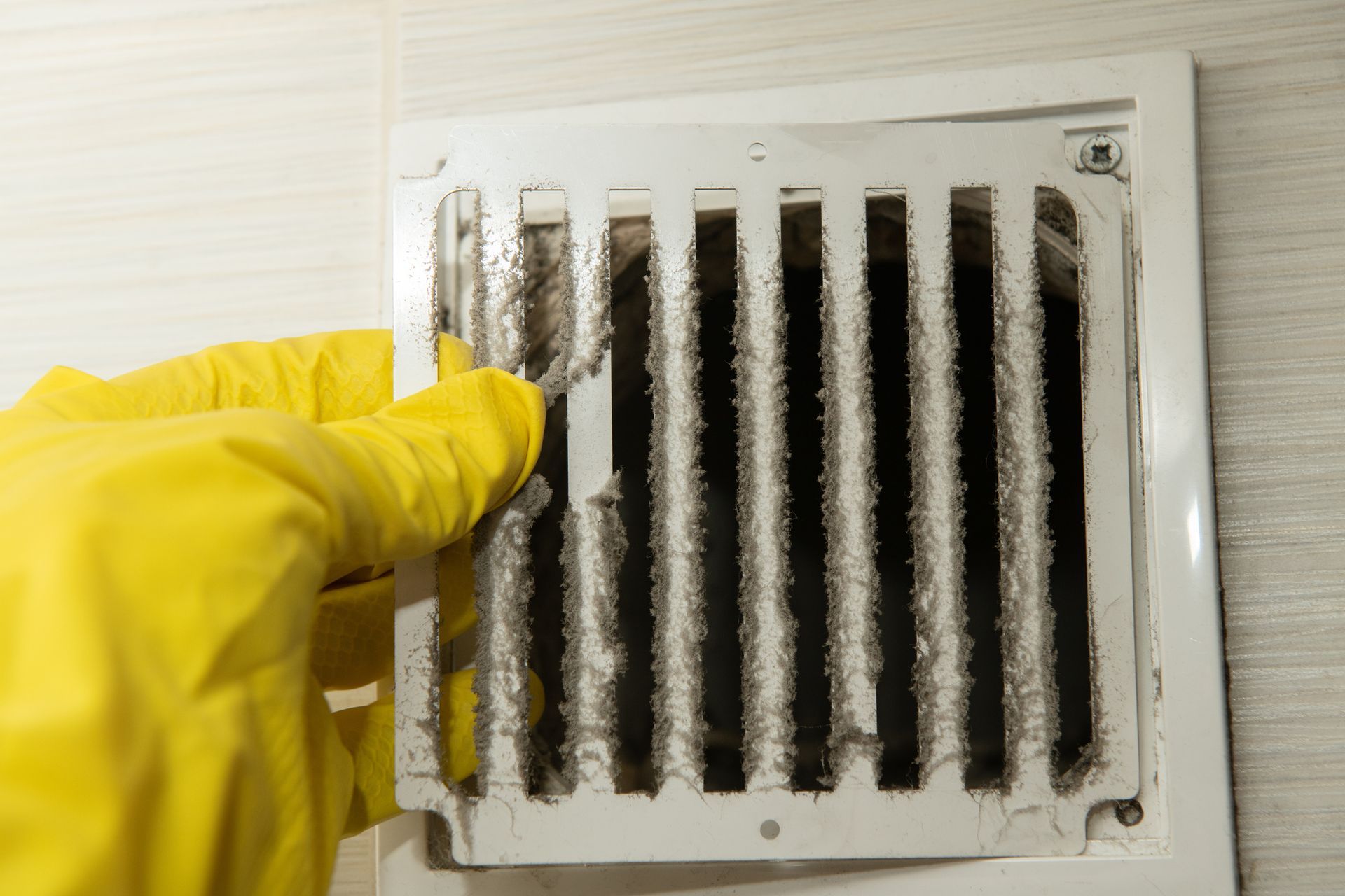 Yellow-gloved hand cleaning a dusty white air vent in a tiled bathroom.