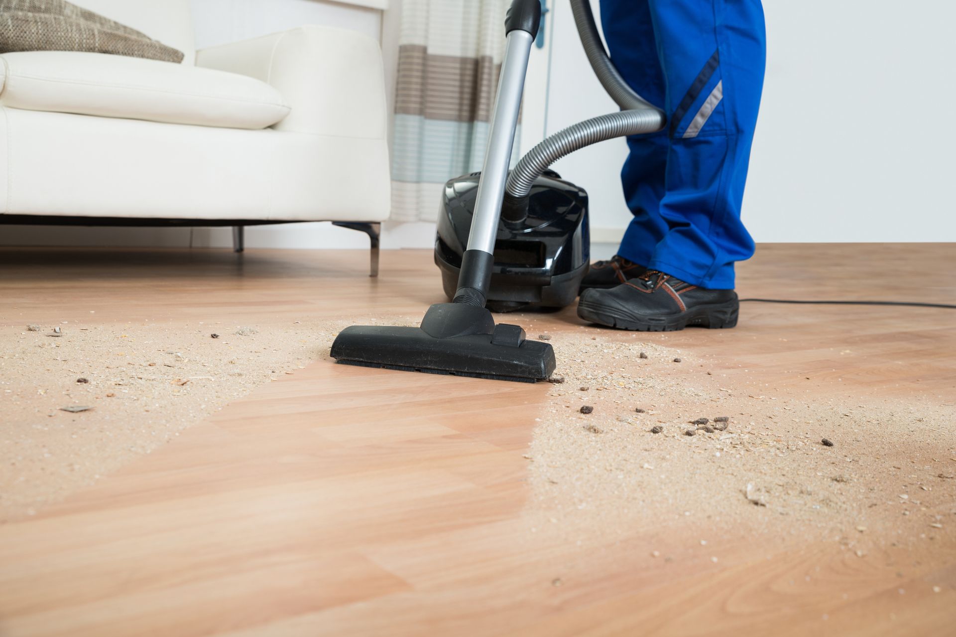 Person in blue coveralls vacuums a dusty wooden floor in a living room.