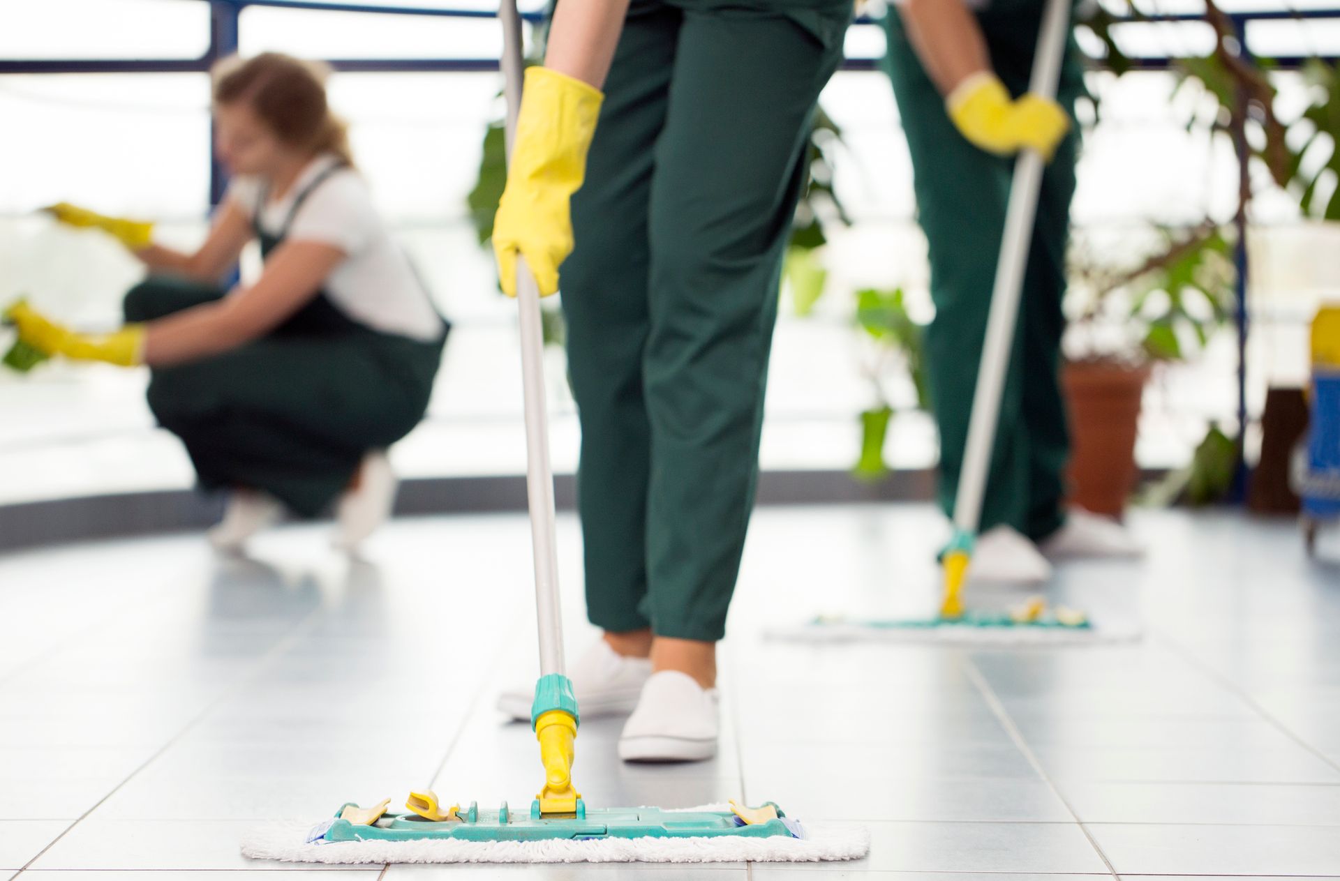 Janitors in green uniforms and yellow gloves mopping a tiled floor in a brightly lit room.