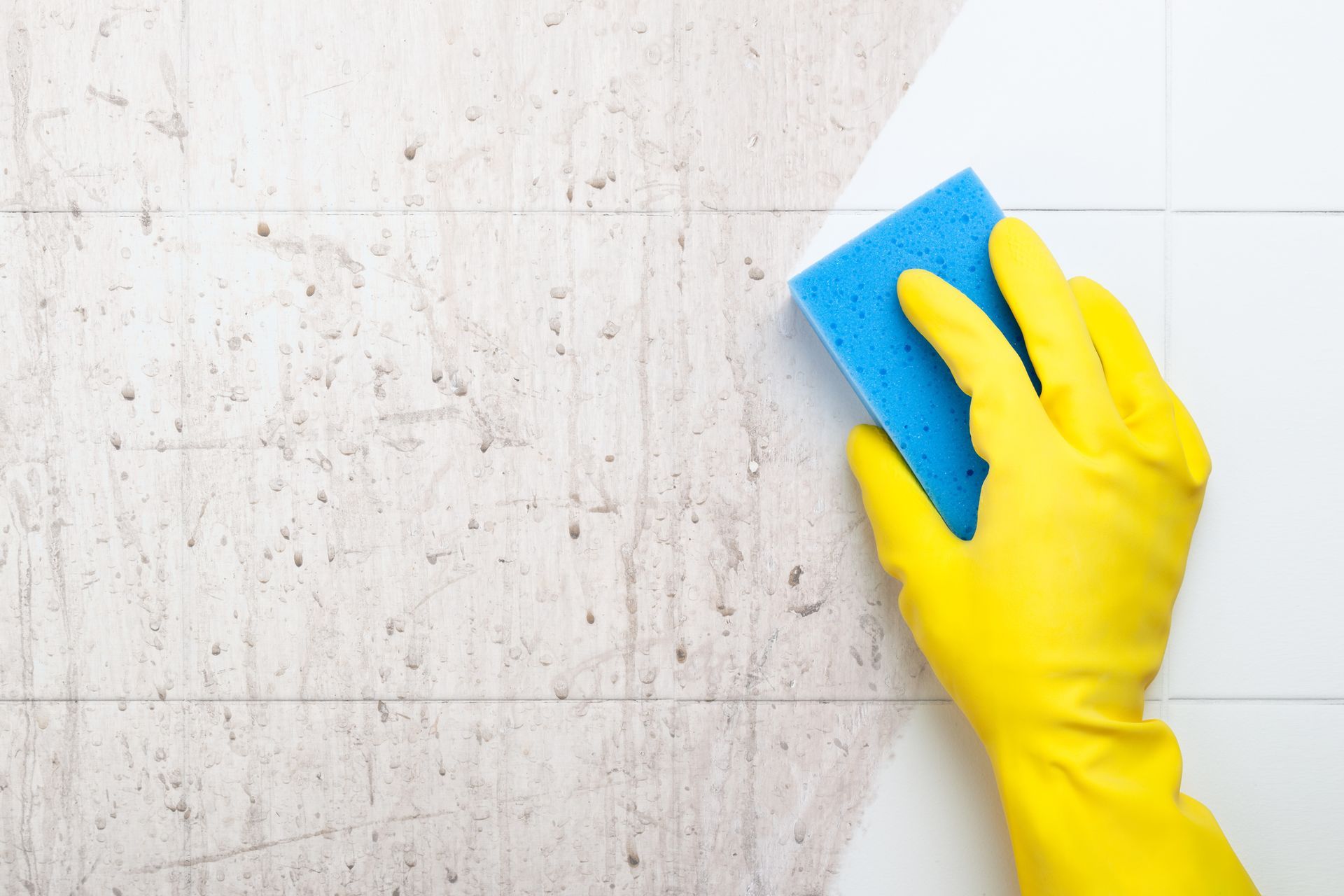 Yellow-gloved hand with blue sponge cleaning tile wall.