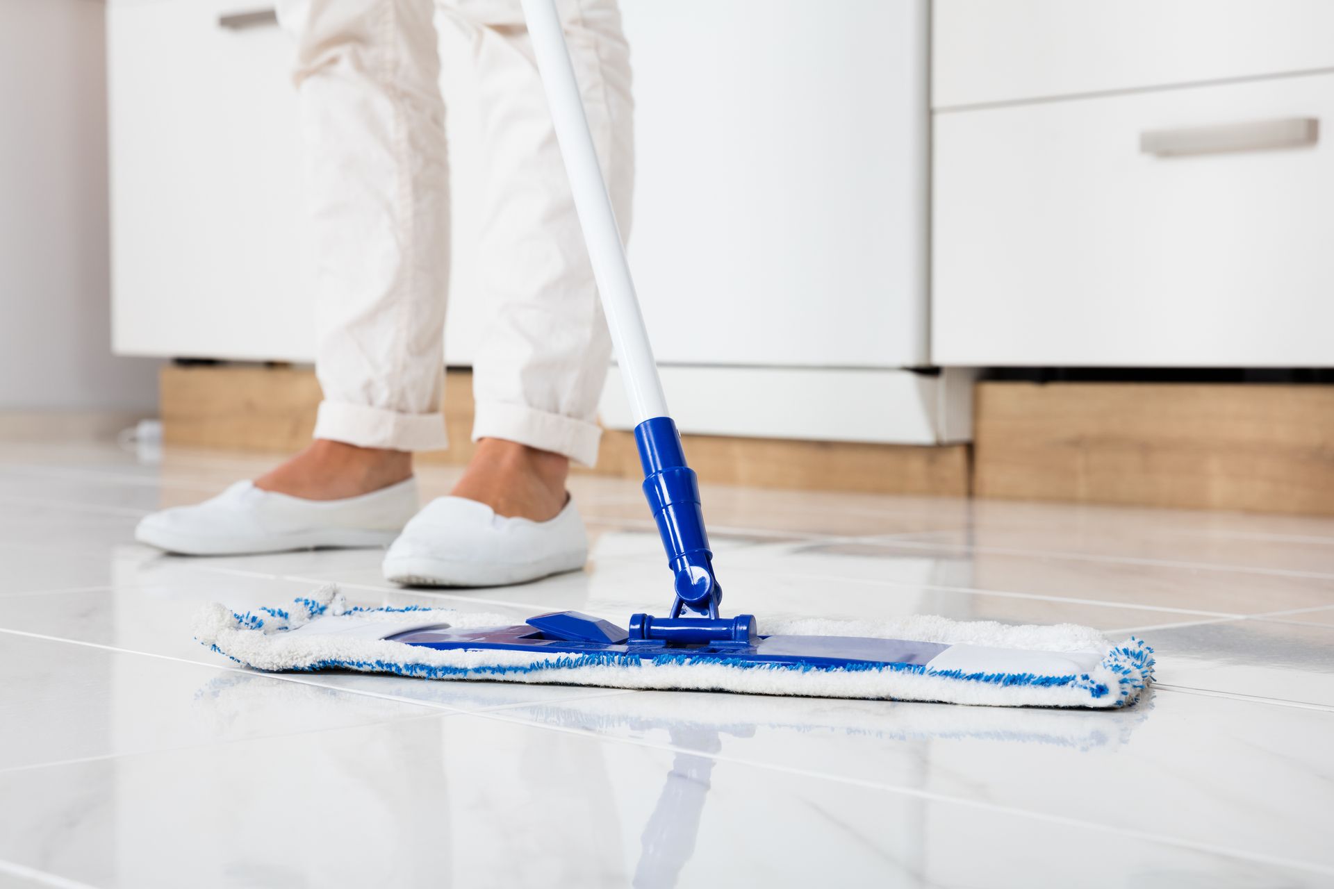 Person mopping a shiny white tile floor in a kitchen, wearing white pants and shoes.