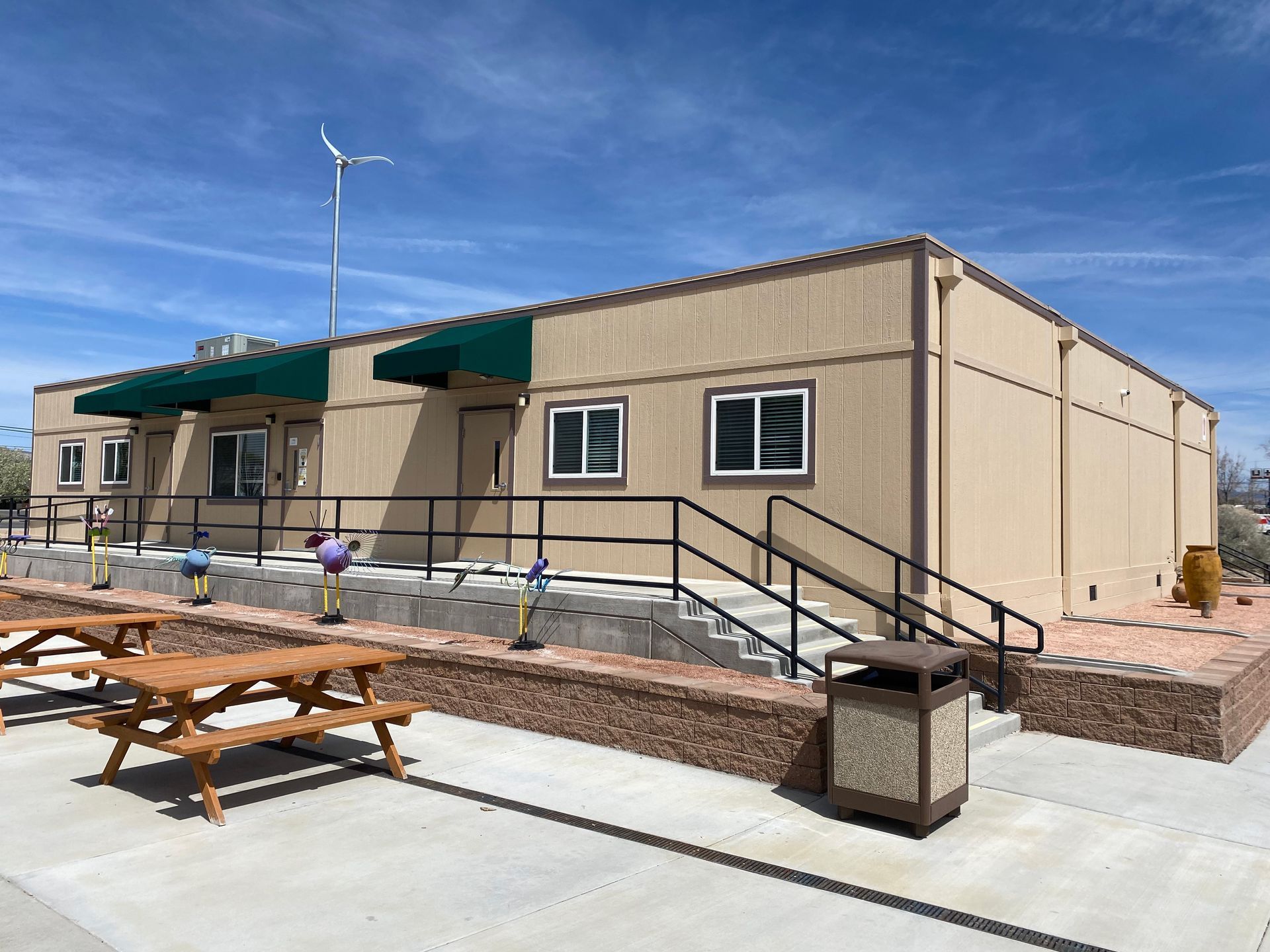 Tan modular building with awning, ramp, picnic tables, and small wind turbine on a sunny day.