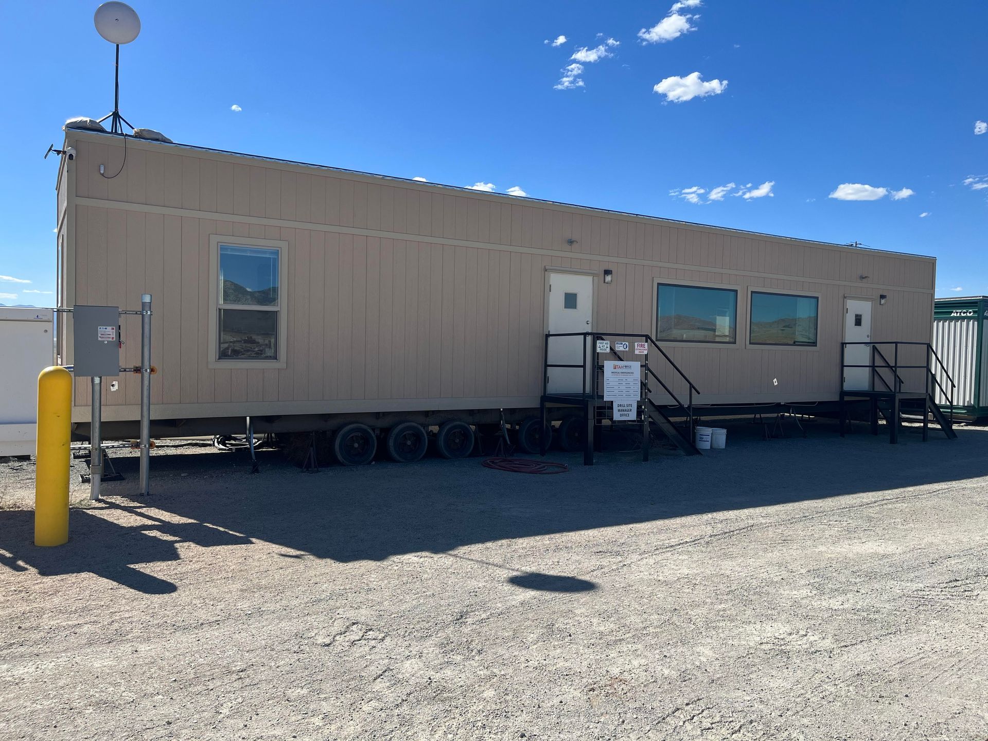 Tan portable office building with antenna, windows, doors, and stairs on gravel under a blue sky.