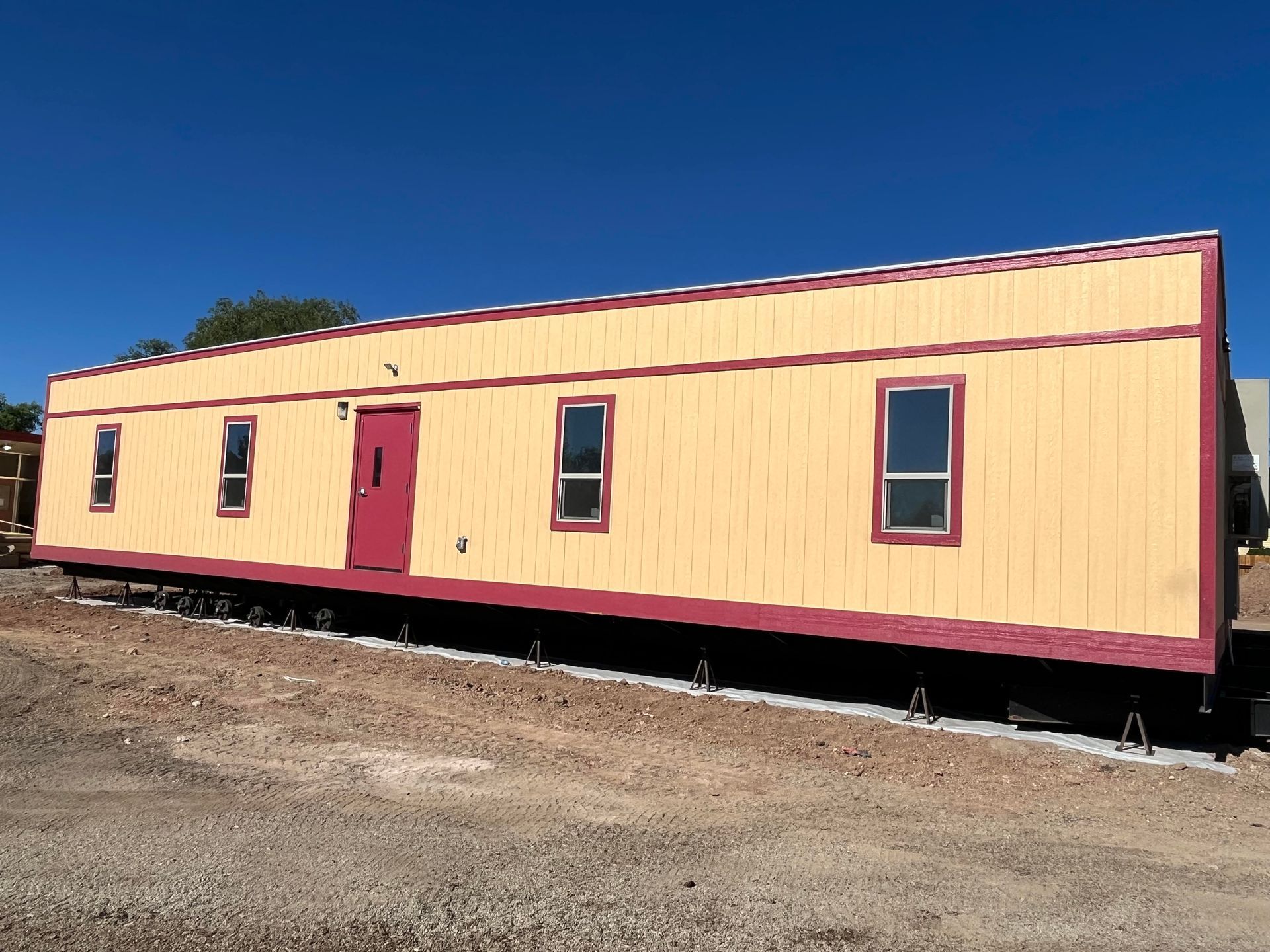 Yellow and red modular building with a red door and windows. Sitting on concrete blocks in a gravel lot.