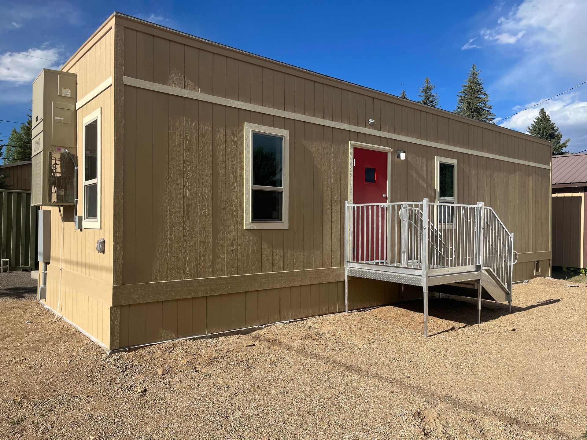 Tan modular building with red door, small porch, and gravel lot.