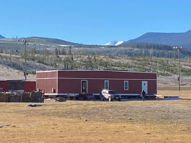 Red rectangular building on wheels, car parked nearby, mountains in the background. Sunny day.