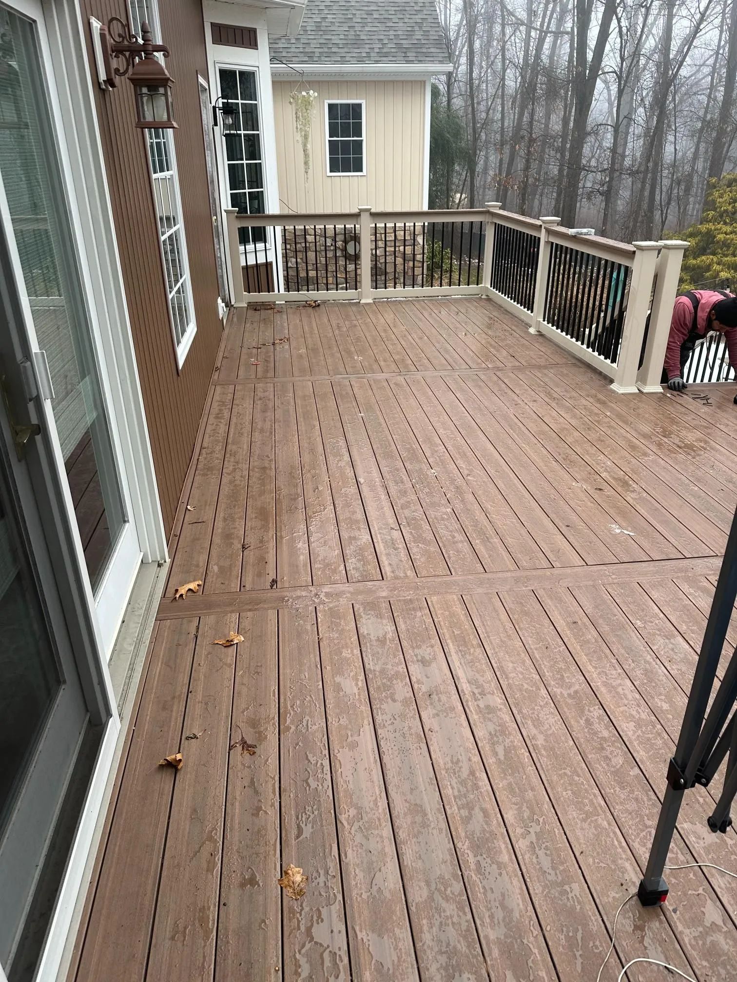 Wet wooden deck with railing beside a house, overlooking a wooded yard and neighboring building.