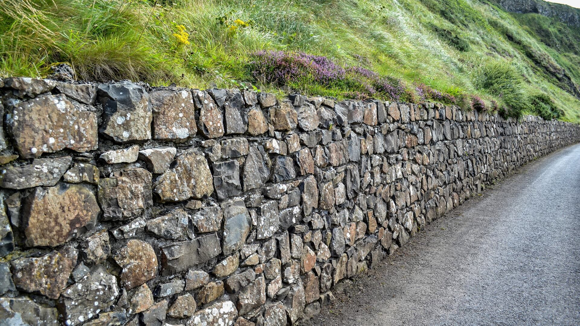A stone retaining wall runs alongside a gravel path with a grassy hill in the background.