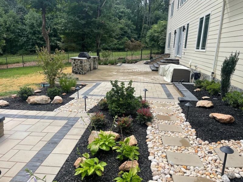 A backyard patio features light pavers with a dark border, stone flower beds, and a stone outdoor grill near a house.