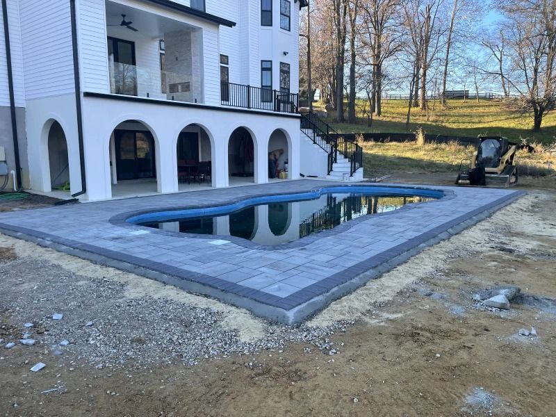 A kidney-shaped swimming pool with gray stone pavers, set beside a multi-story house with white arches and a patio.