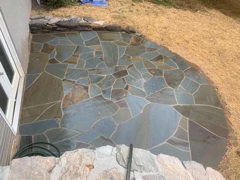 A flagstone patio made of irregularly shaped, blue-gray stones next to a house with a stone retaining wall.