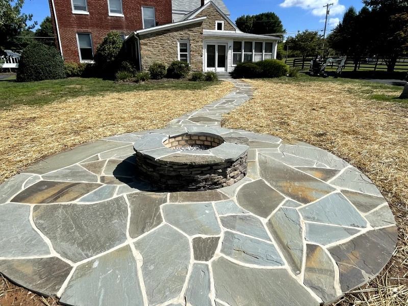 A stone fire pit sits in the center of a circular flagstone patio, connected by a stone path to a house in the background.