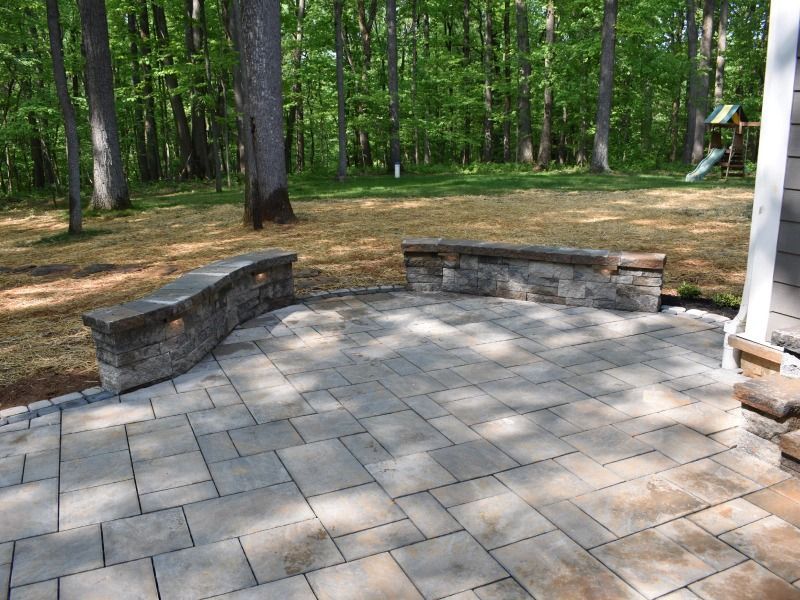 A stone patio with two curved, built-in stone benches overlooking a backyard with trees and a wooden play set.
