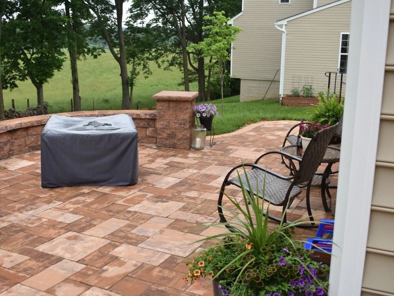 A stone patio with a covered fire pit, outdoor chairs, potted flowers, and a scenic grassy field in the background.