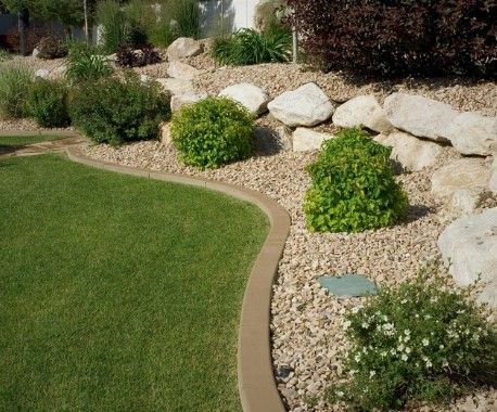 A curved concrete border separates a vibrant green lawn from a rocky landscape featuring small shrubs and large boulders.