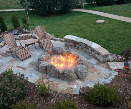 A stone-rimmed fire pit glows in a backyard patio with two wooden chairs and a curved stone seating wall on a grassy lawn.
