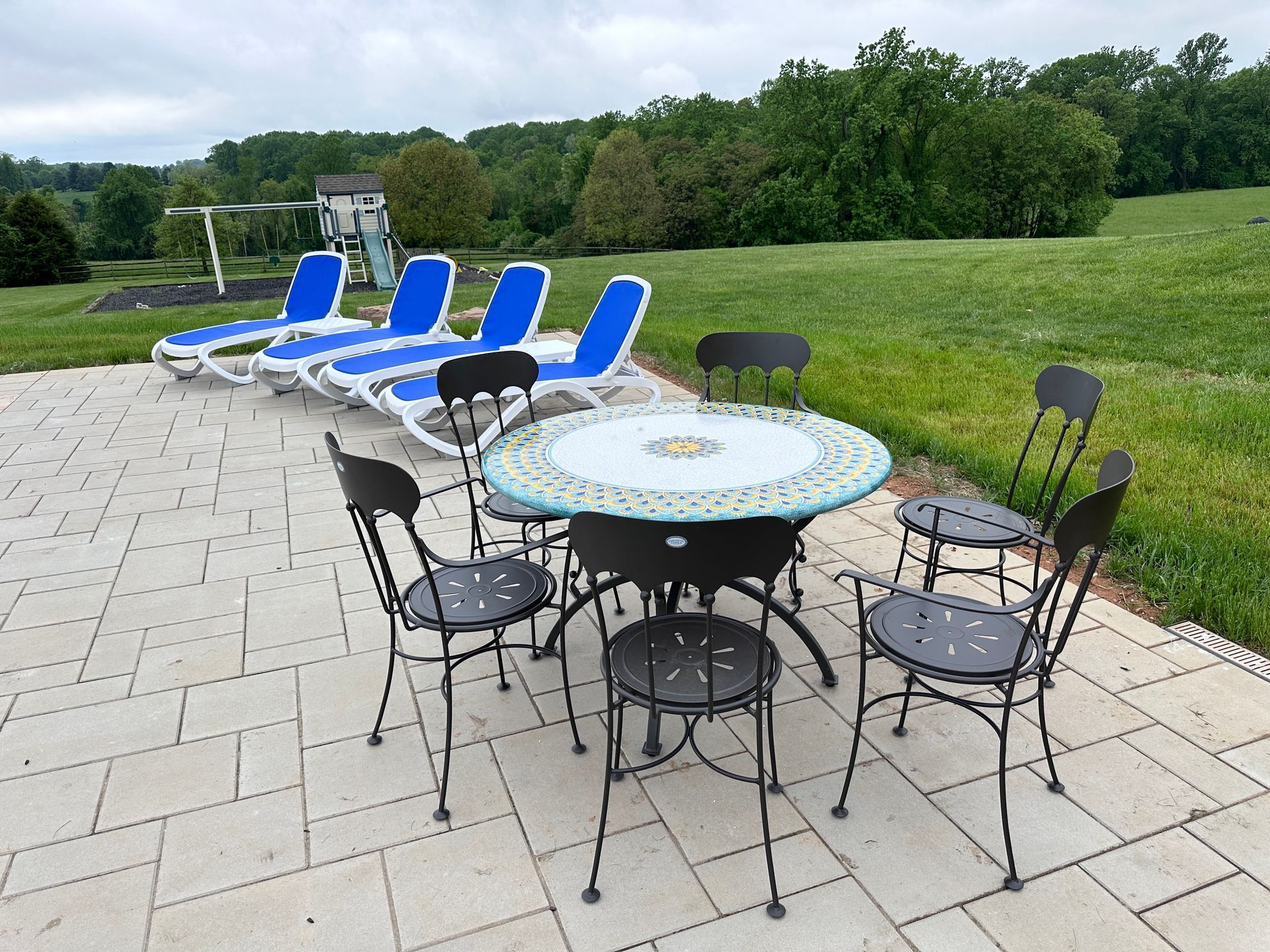 A patio set with a round tiled table and chairs sits on stone pavers beside four blue lounge chairs in a grassy yard.
