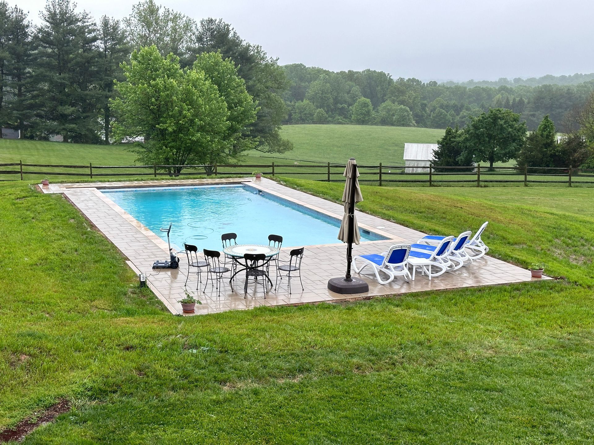 A rectangular swimming pool with patio furniture on a concrete deck, surrounded by a grassy lawn and a rural landscape.