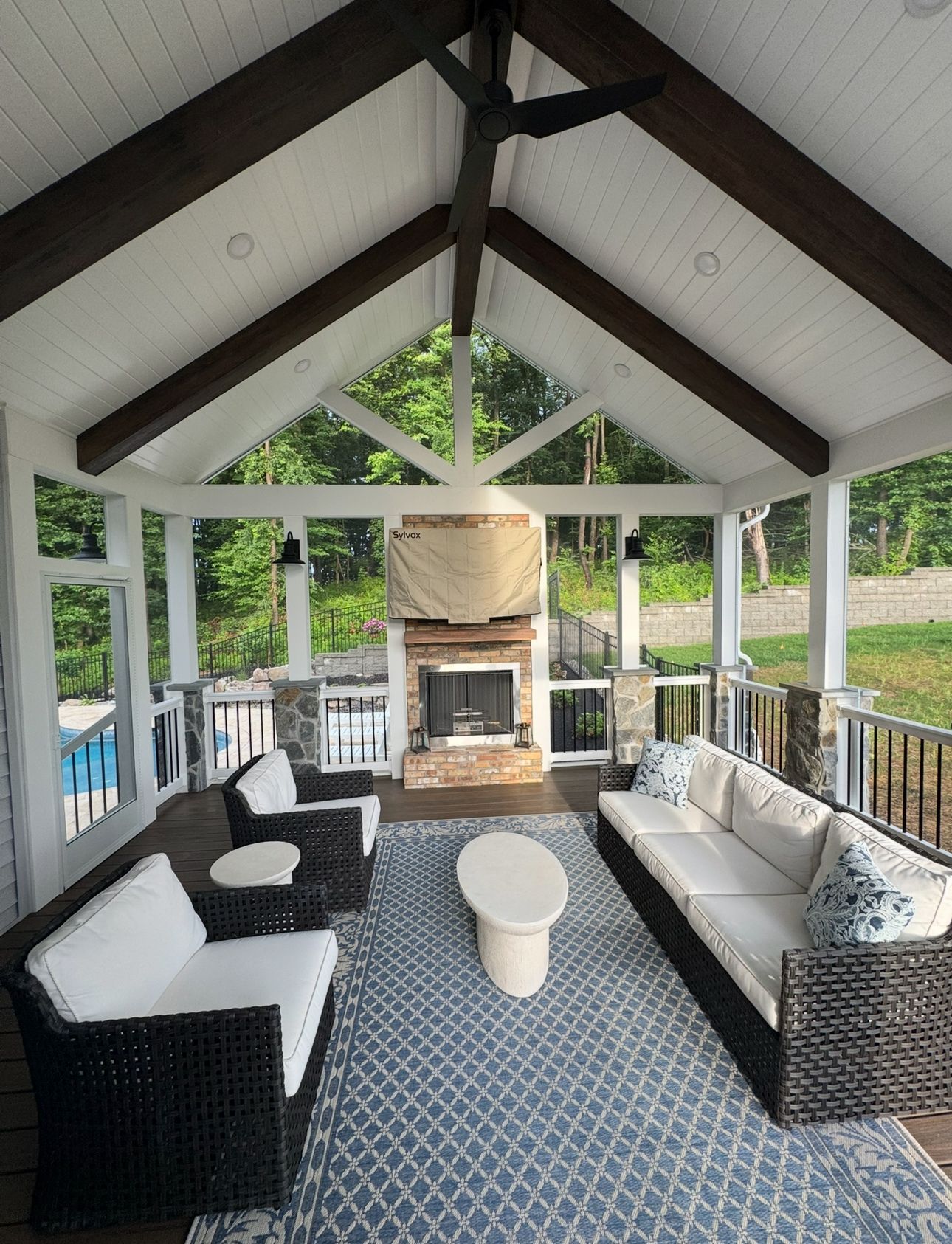 A bright, covered porch with a fireplace, comfortable outdoor furniture, and a blue patterned rug overlooking a yard.