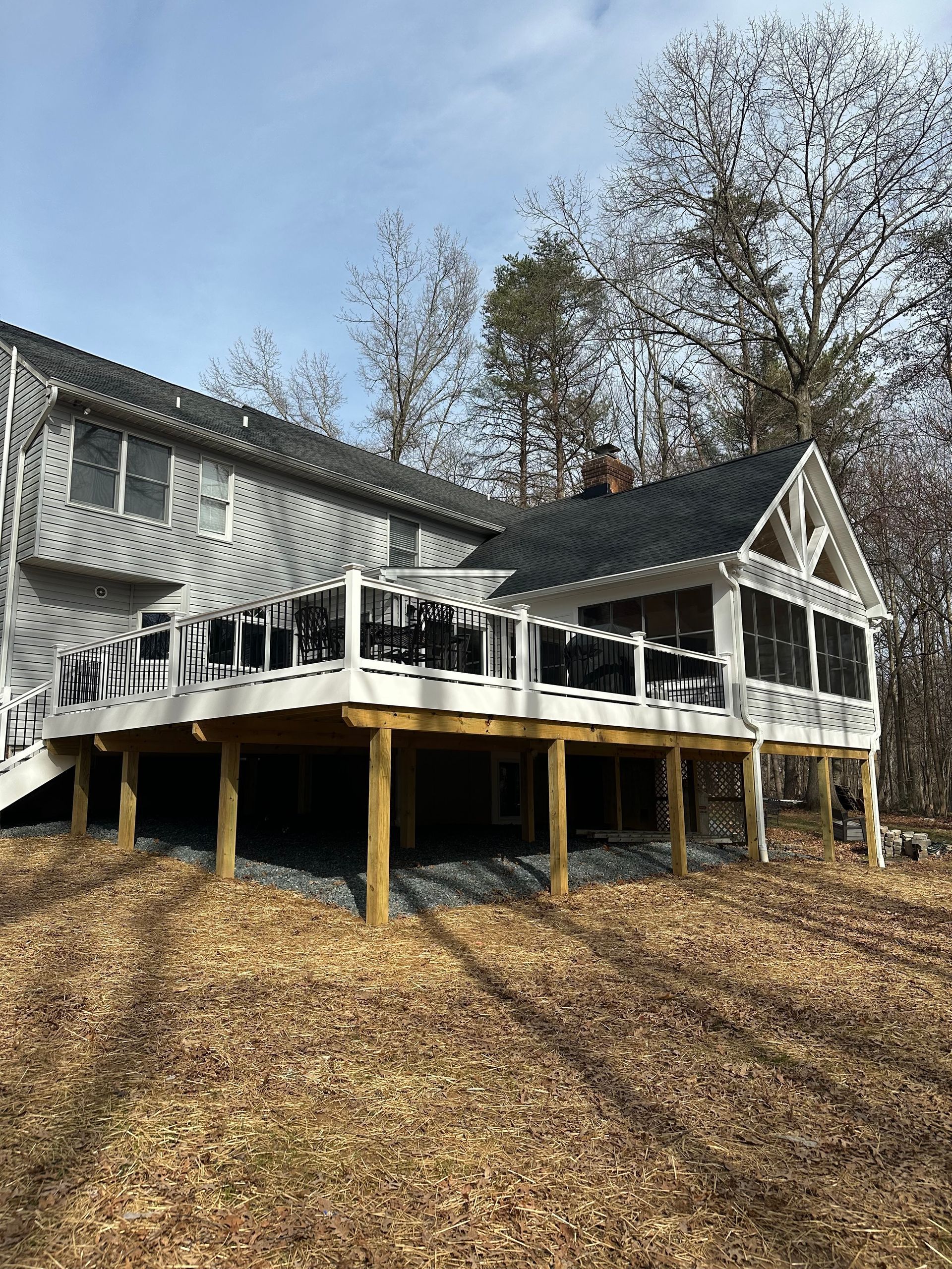 A large, elevated white deck with a black railing attached to a two-story gray house in a wooded area.