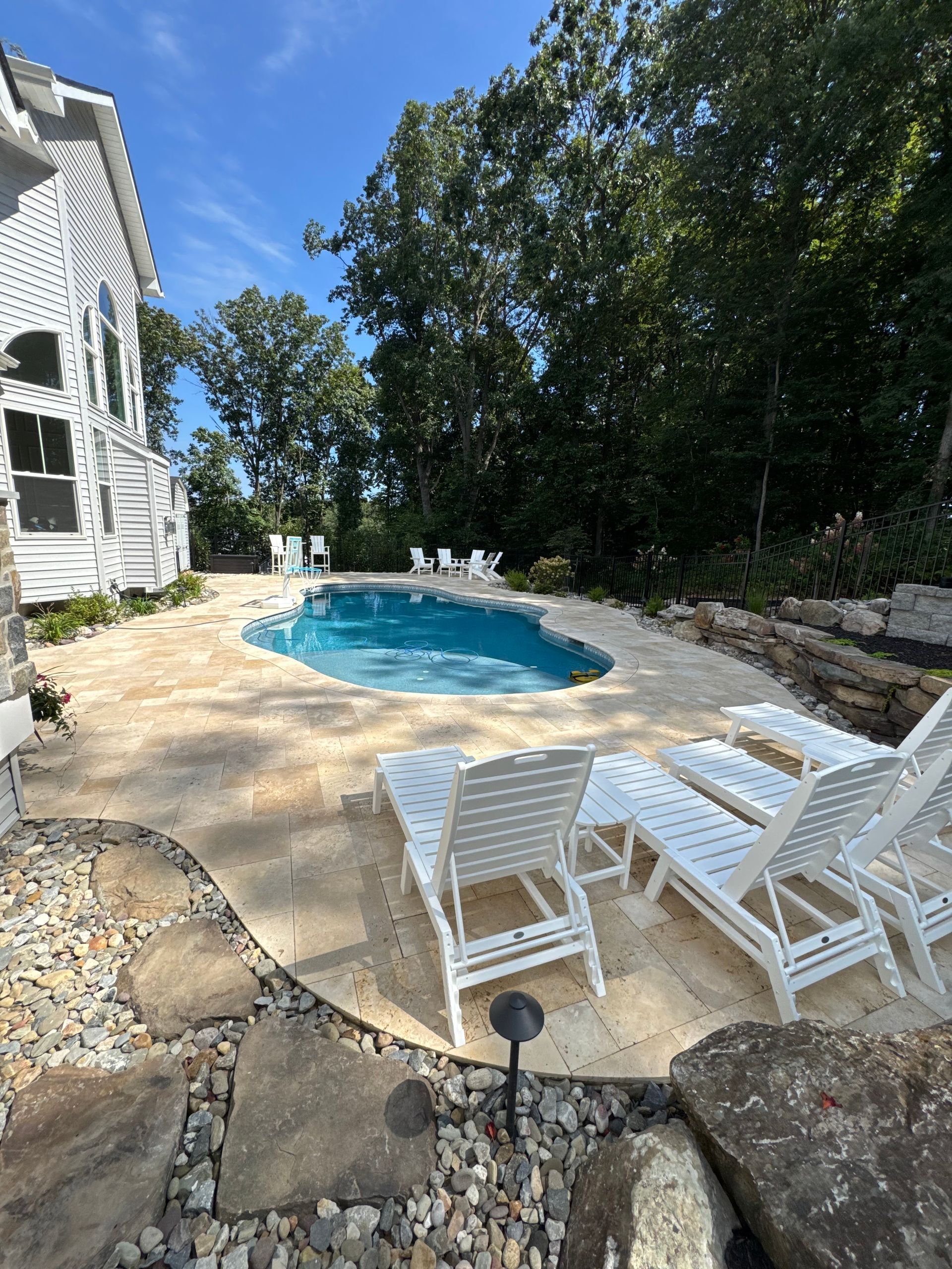 A backyard swimming pool with lounge chairs on a stone patio, next to a white house surrounded by trees under a blue sky.