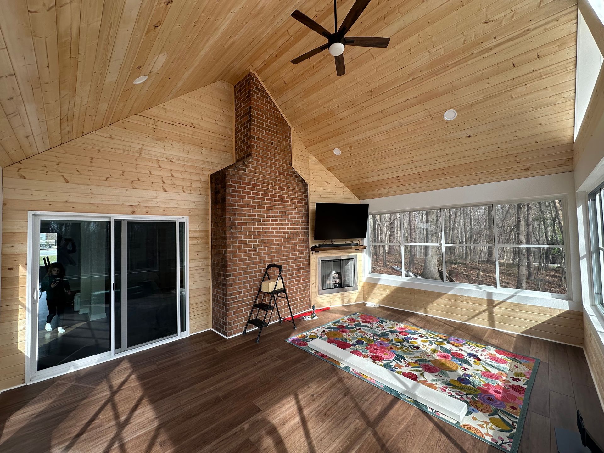 A sunroom with wood-paneled walls and ceiling, featuring a brick fireplace, sliding glass door, and a colorful rug.