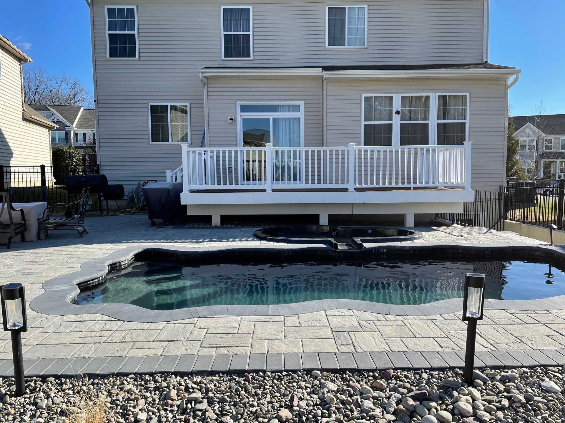 A tan two-story house with a white deck, a custom inground pool with stone coping, and a paver patio in the backyard.