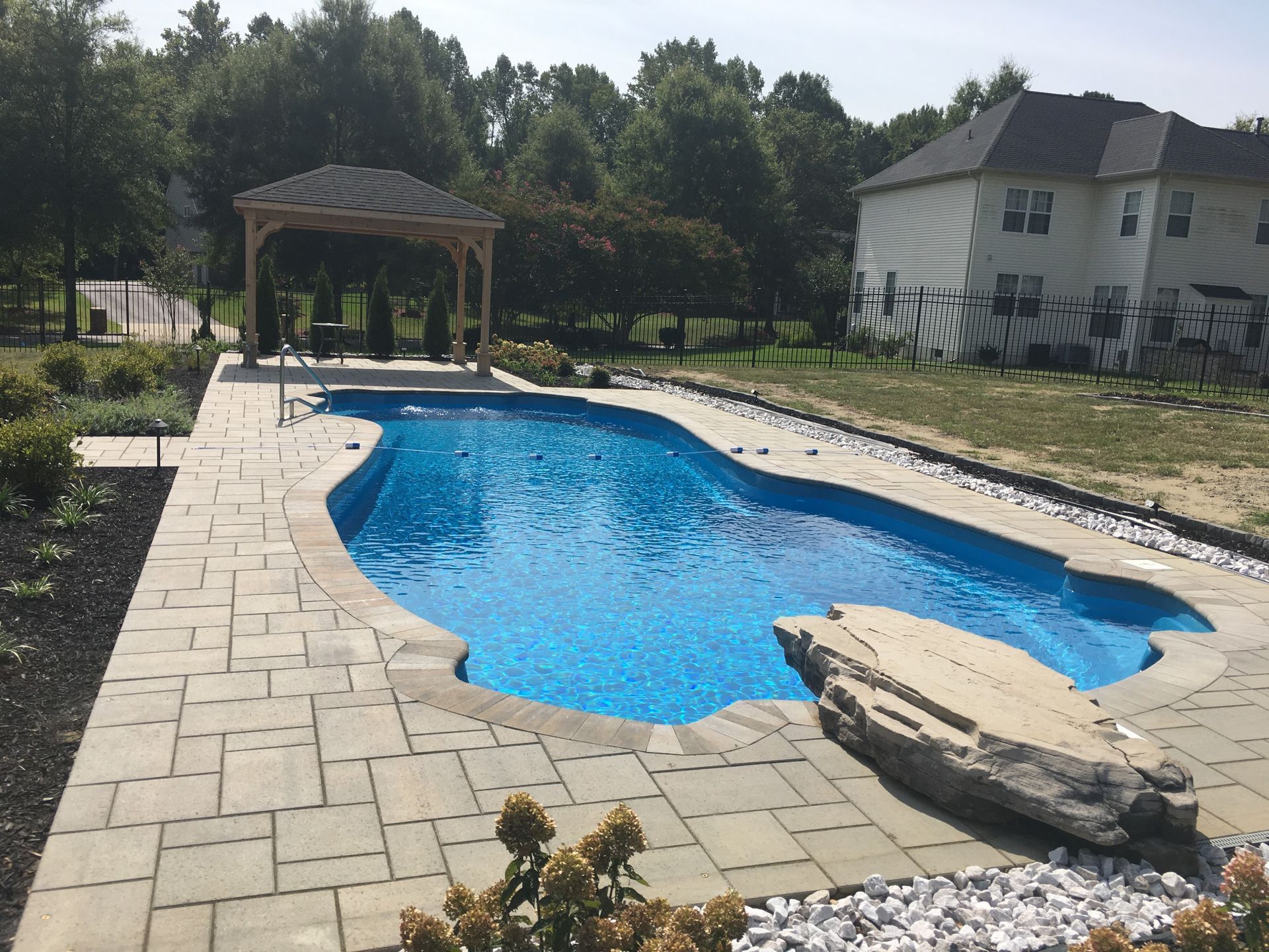 A backyard swimming pool with a stone patio, a gazebo at the far end, and a large boulder at the edge of the pool.