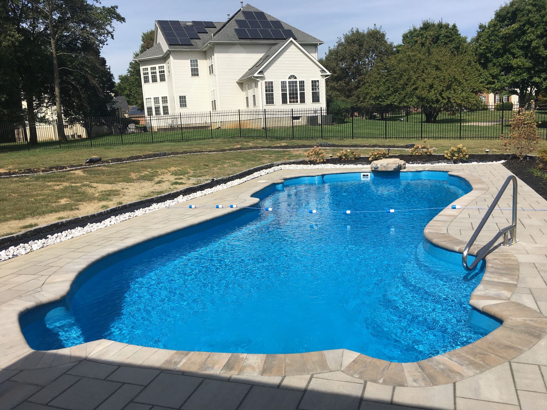A bright blue swimming pool with a stone patio surround in front of a white house with solar panels on the roof.