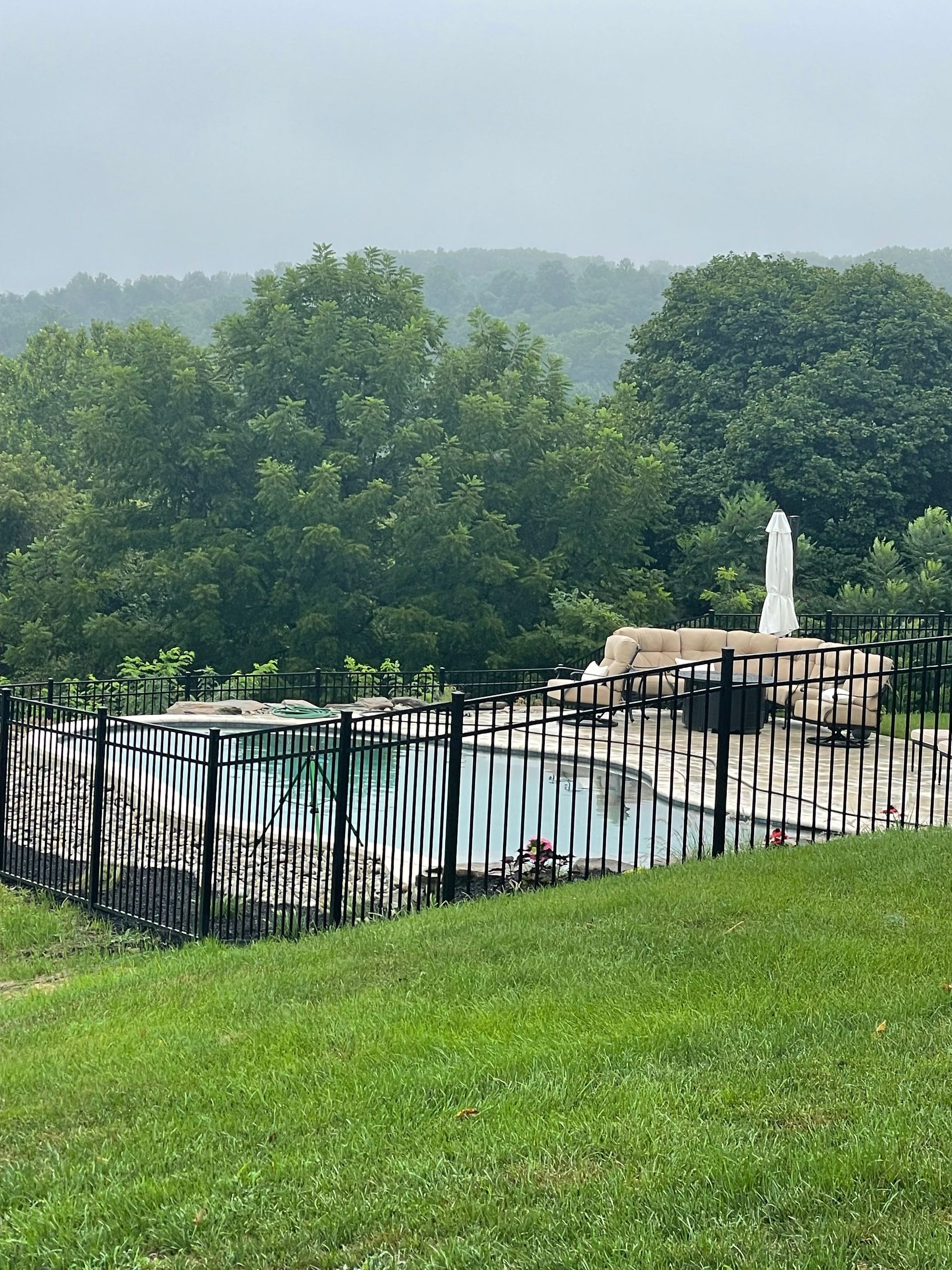 A dark metal fence encloses a backyard swimming pool with a stone waterfall feature, set against a backdrop of trees.