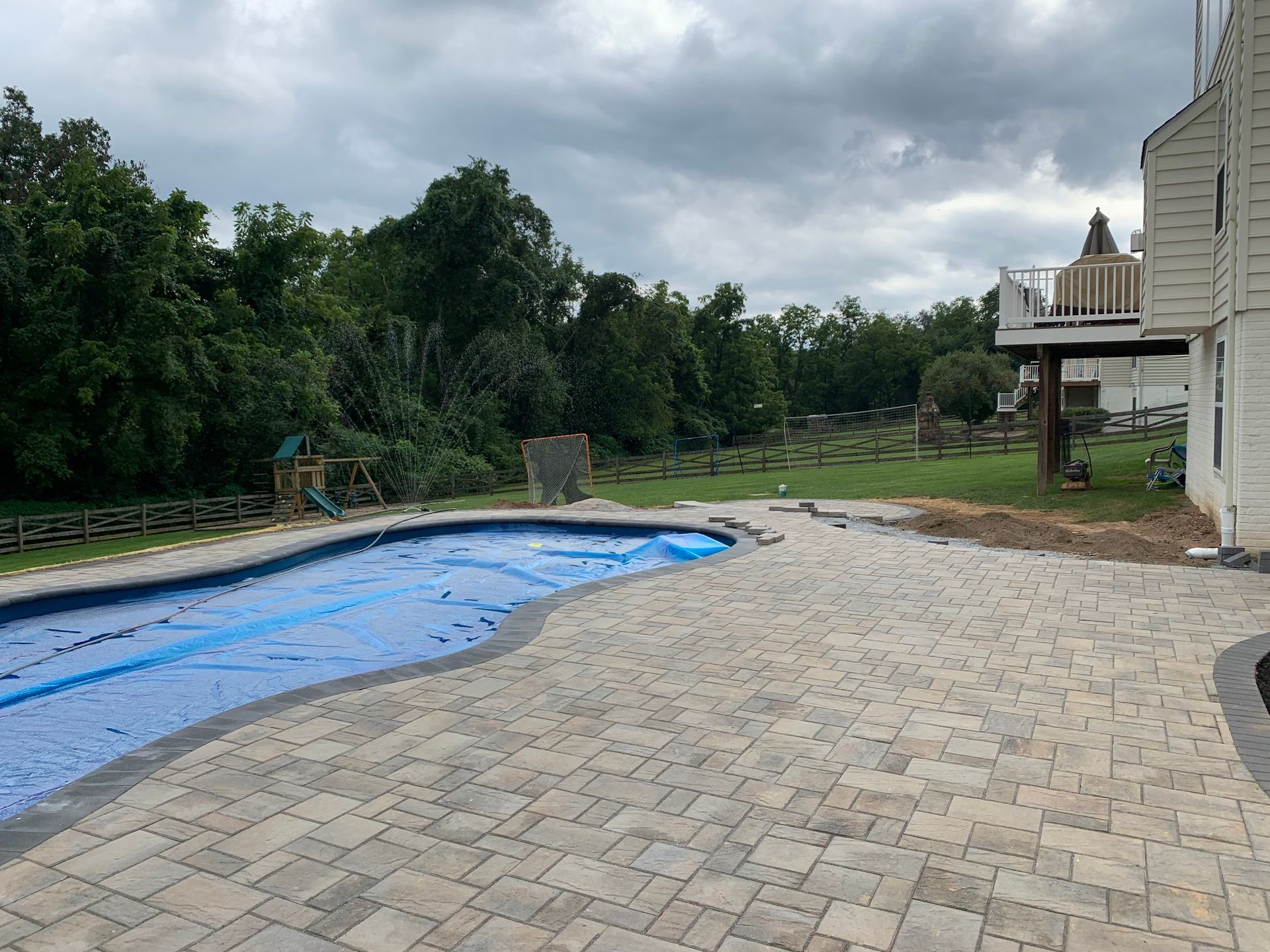 A blue pool cover on a curved pool beside a light-colored paver patio with a house and trees in the background.