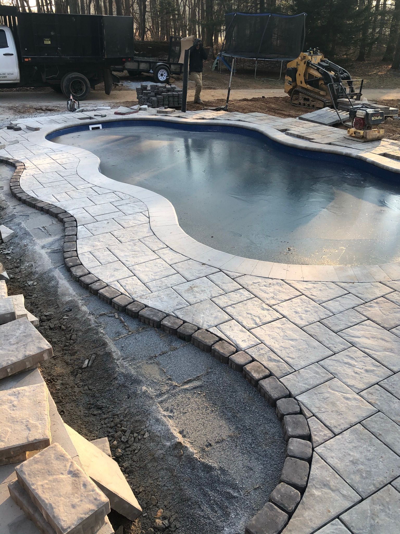 An unfinished swimming pool patio under construction, showing grey pavers partially laid around the curved pool edge.