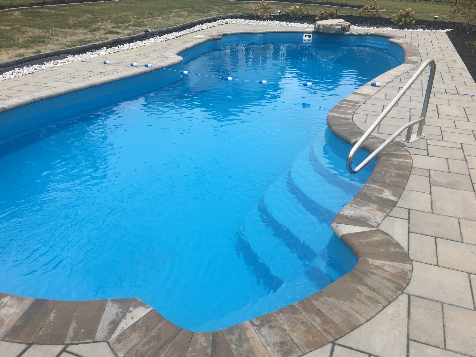 A bright blue inground swimming pool with curved steps, a metal handrail, and surrounding tan stone patio pavers.