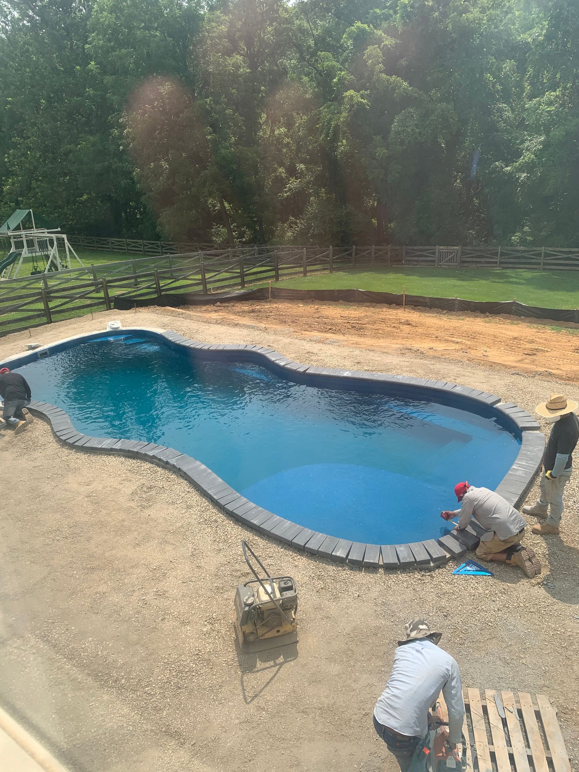 Three workers install stone coping around the edge of a curved, blue backyard swimming pool under construction.
