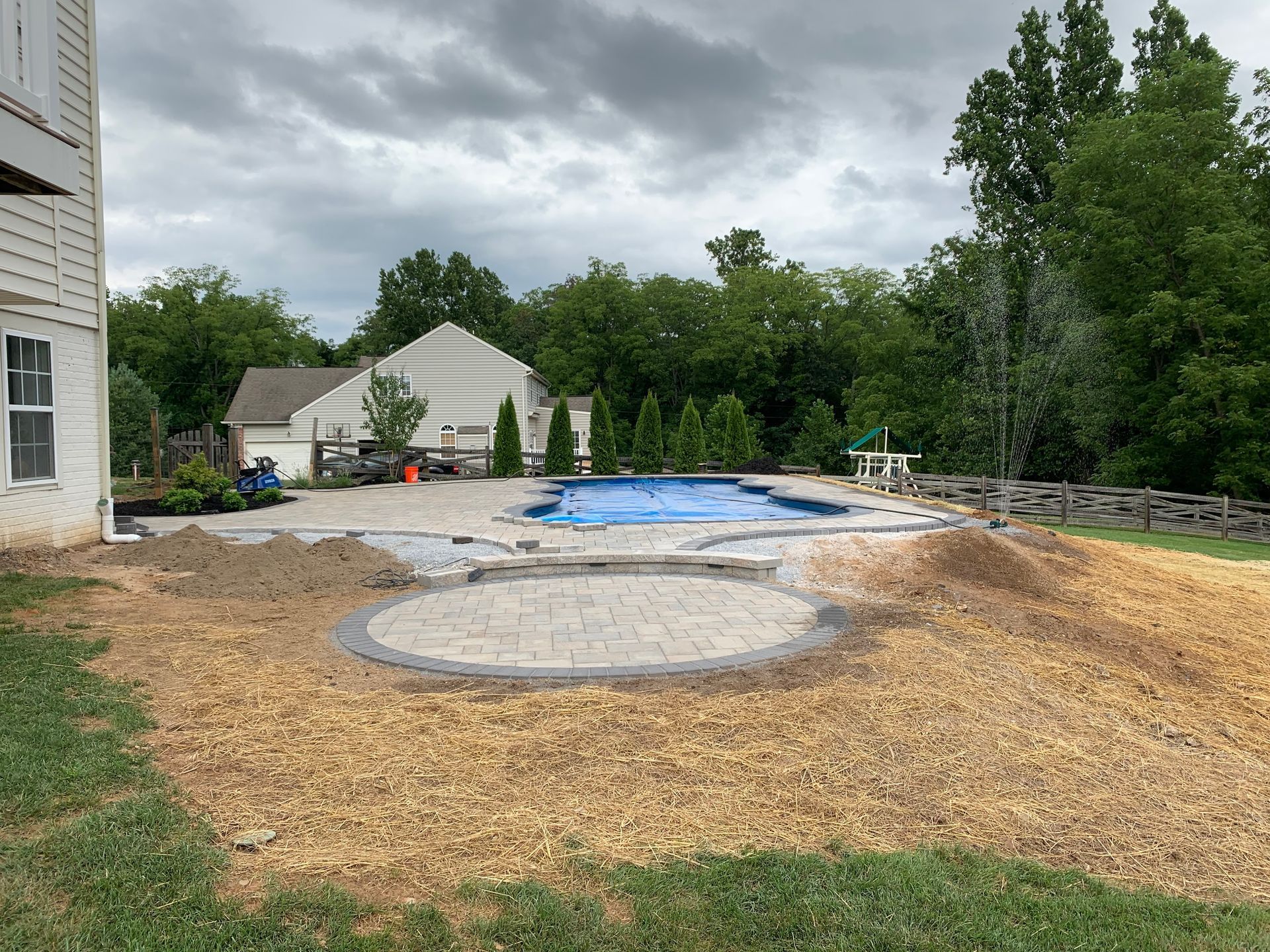 A construction site showing a backyard pool with a blue cover, a circular stone patio, and unfinished landscaping.