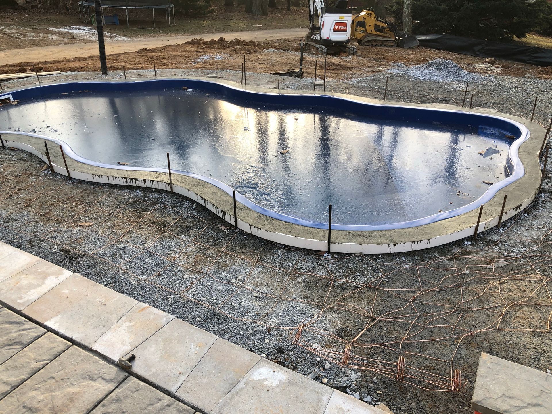 An irregularly shaped swimming pool under construction, surrounded by gravel, wooden forms, and a paved stone walkway.