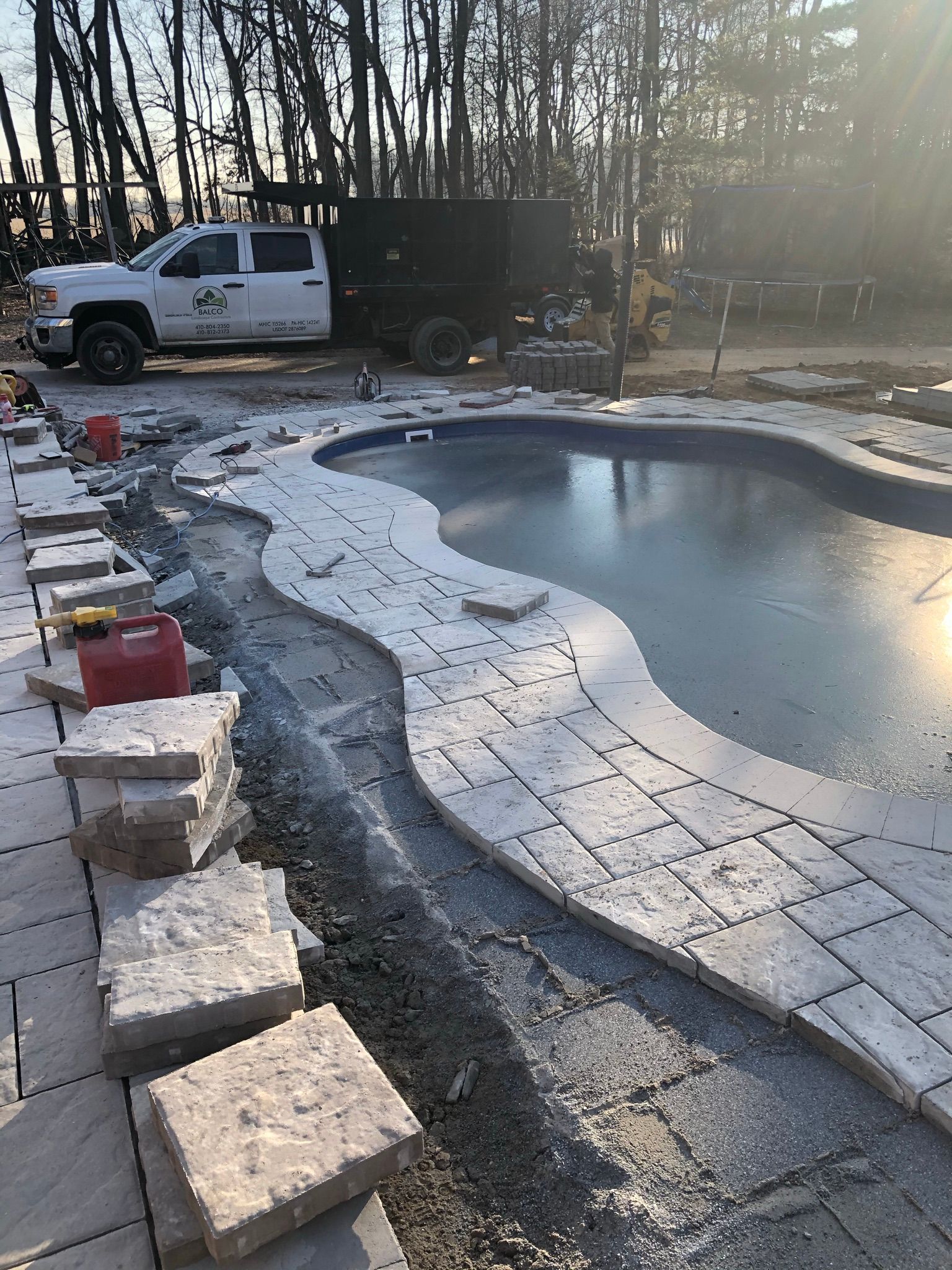A white truck parked next to a swimming pool under construction with light-colored pavers laid along the curved edge.
