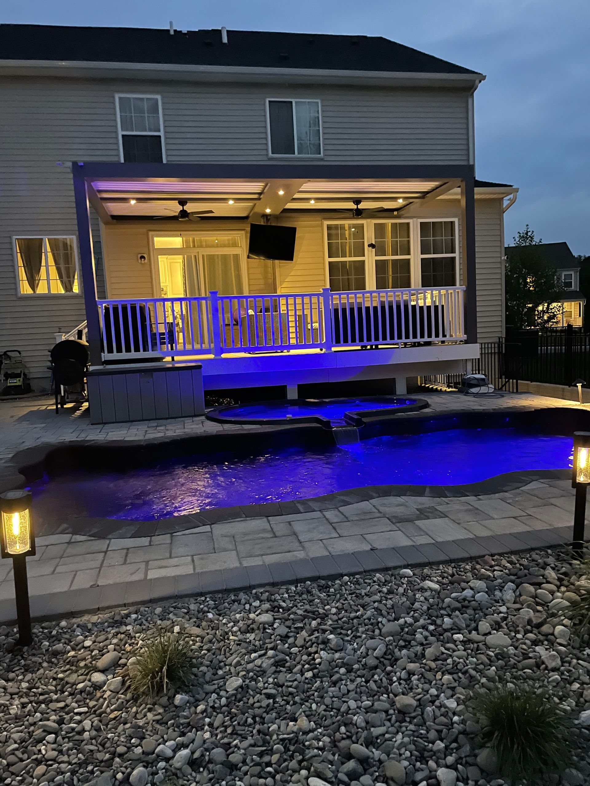 A backyard patio with a white railing, glowing blue lights, and an illuminated swimming pool at dusk.