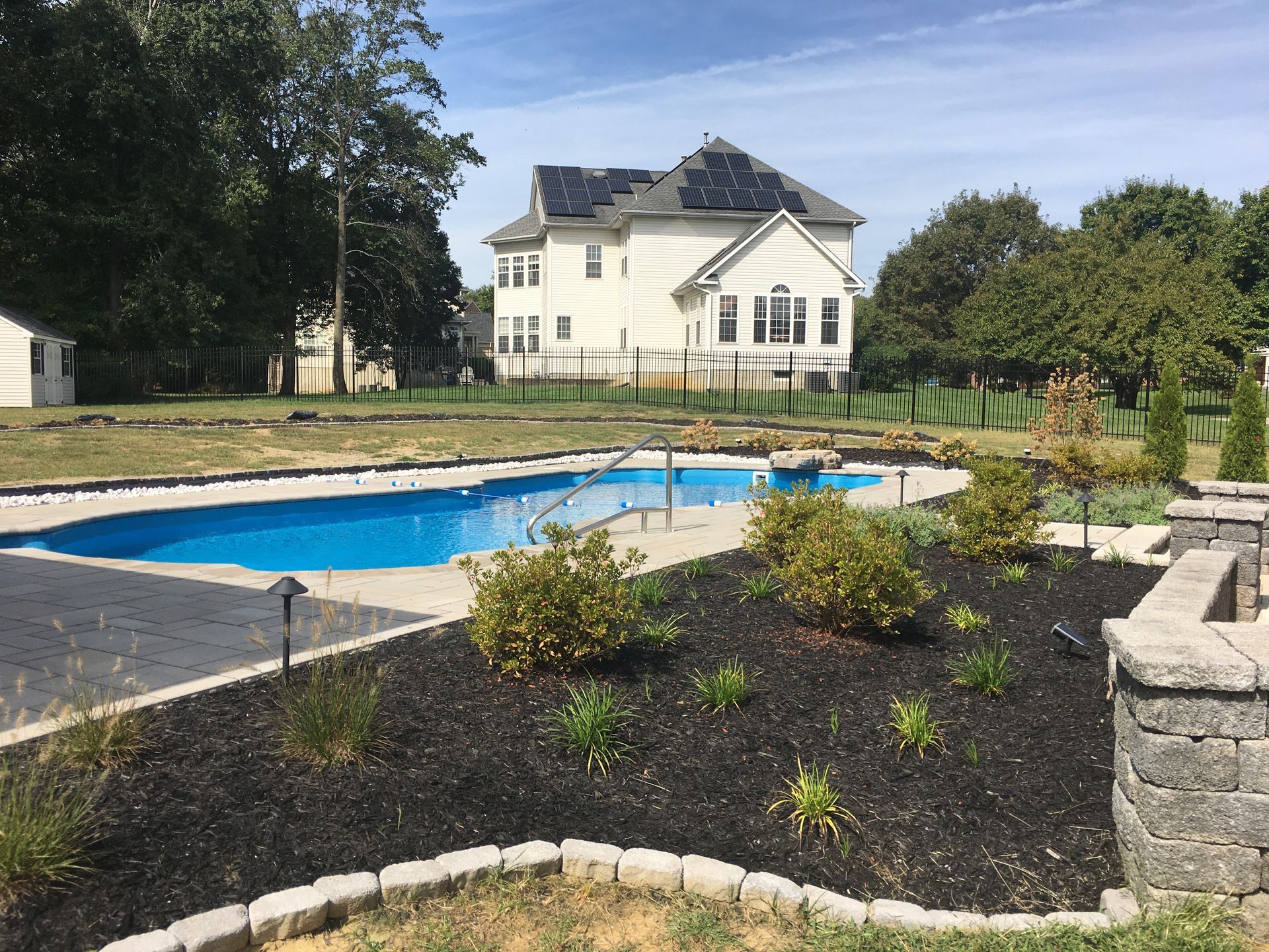 A backyard swimming pool with a stone retaining wall and mulch bed in the foreground, and a large house in the distance.