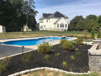 A swimming pool patio with landscaped mulch beds bordered by grey stones, leading toward a small white shed in the yard.