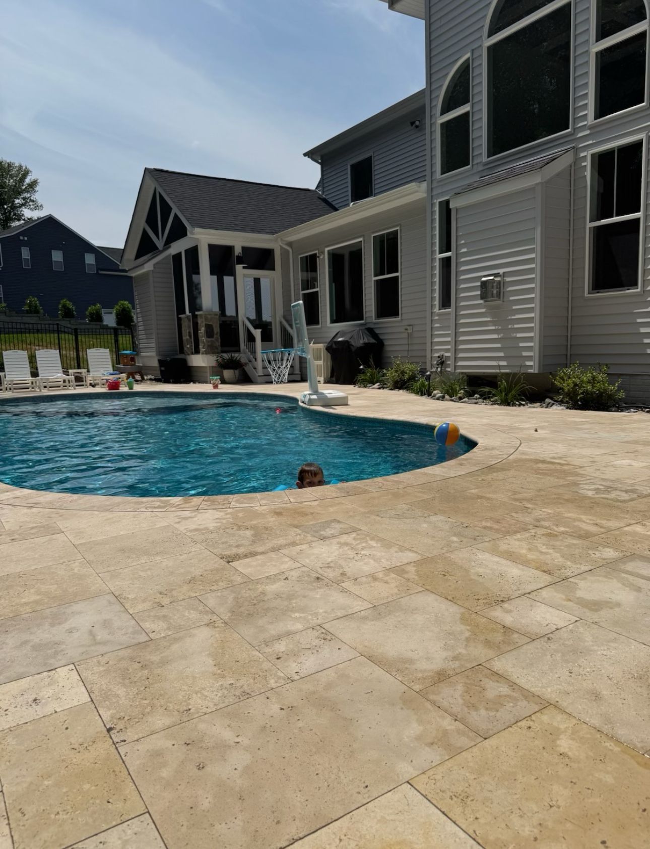 A person swimming in a backyard pool next to a light-colored stone patio and a two-story gray house.