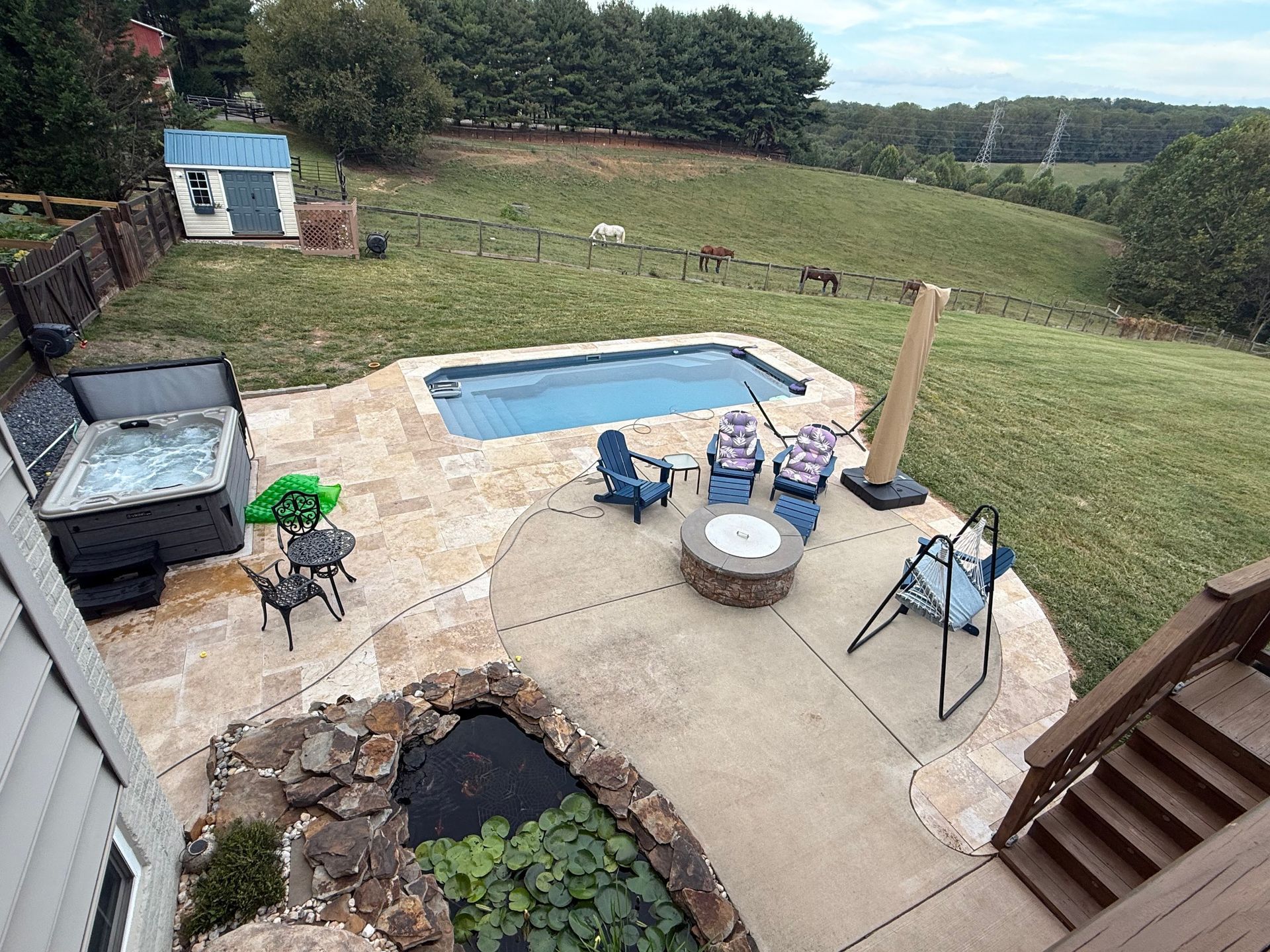 A small backyard pond with a stone waterfall, surrounded by rocks, lush green plants, and potted flowers.