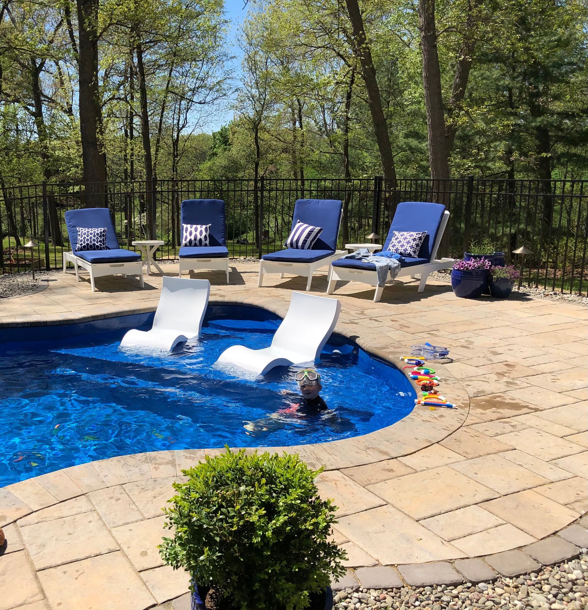 A child plays in a blue pool with submerged lounge chairs, surrounded by patio seating, trees, and a black fence.