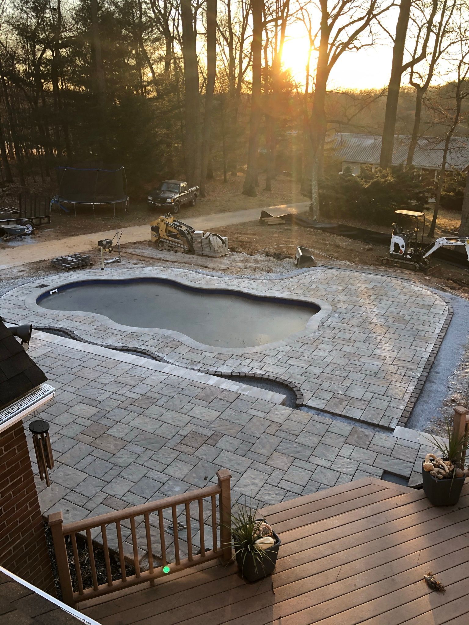 A backyard with a newly installed stone paver patio surrounding a swimming pool at sunset, viewed from a wooden deck.