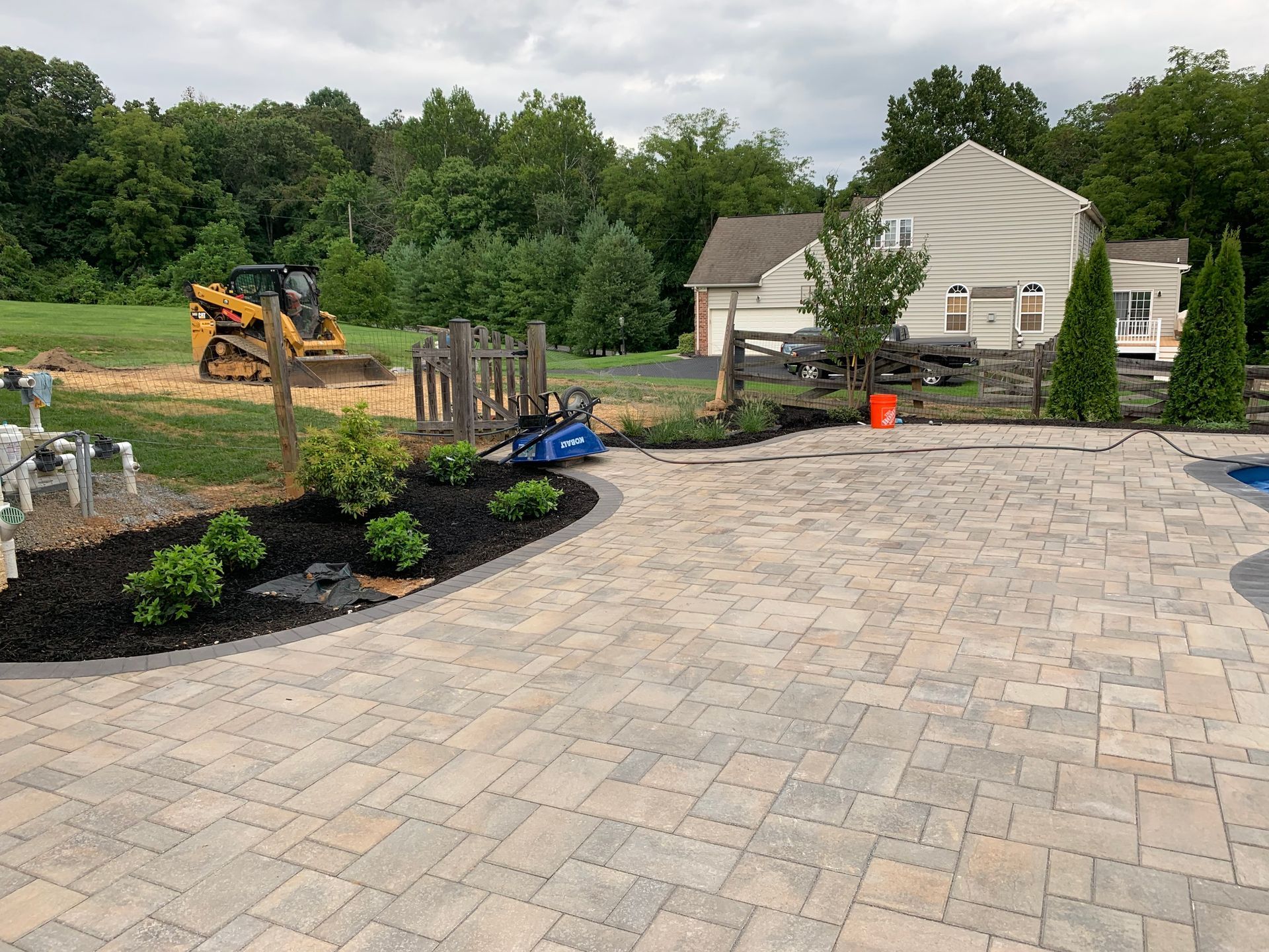 A backyard patio featuring light-colored pavers, a landscaped mulched bed with shrubs, and construction equipment nearby.