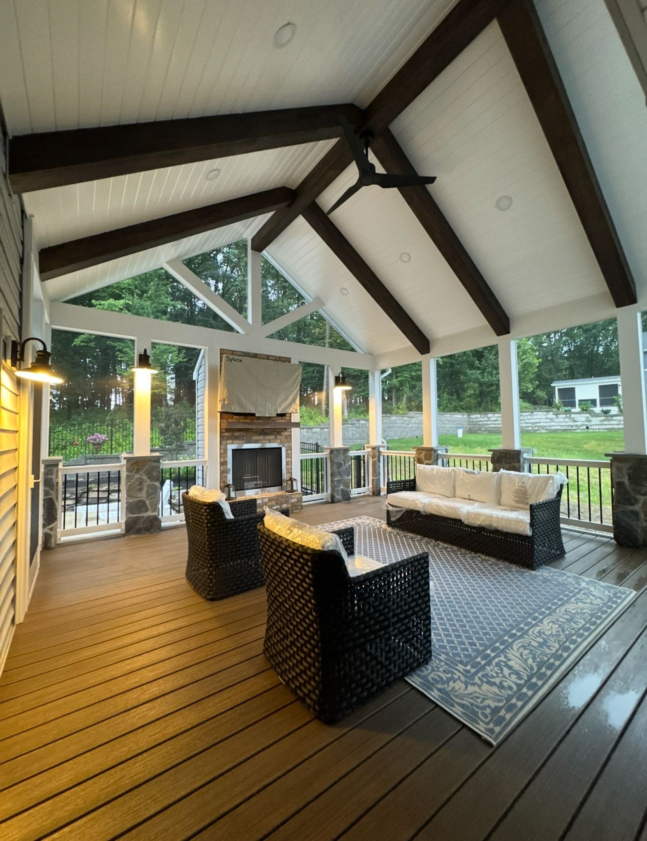 A covered patio featuring a stone fireplace, dark wicker furniture, a patterned rug, and exposed wooden ceiling beams.