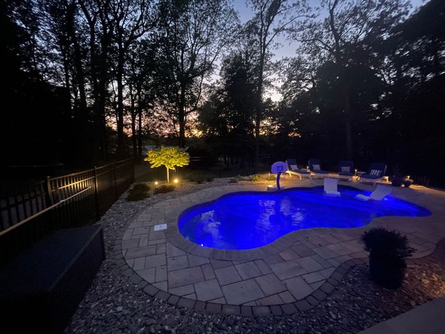 A blue-lit backyard swimming pool with a stone patio at dusk, surrounded by trees and landscape lighting.
