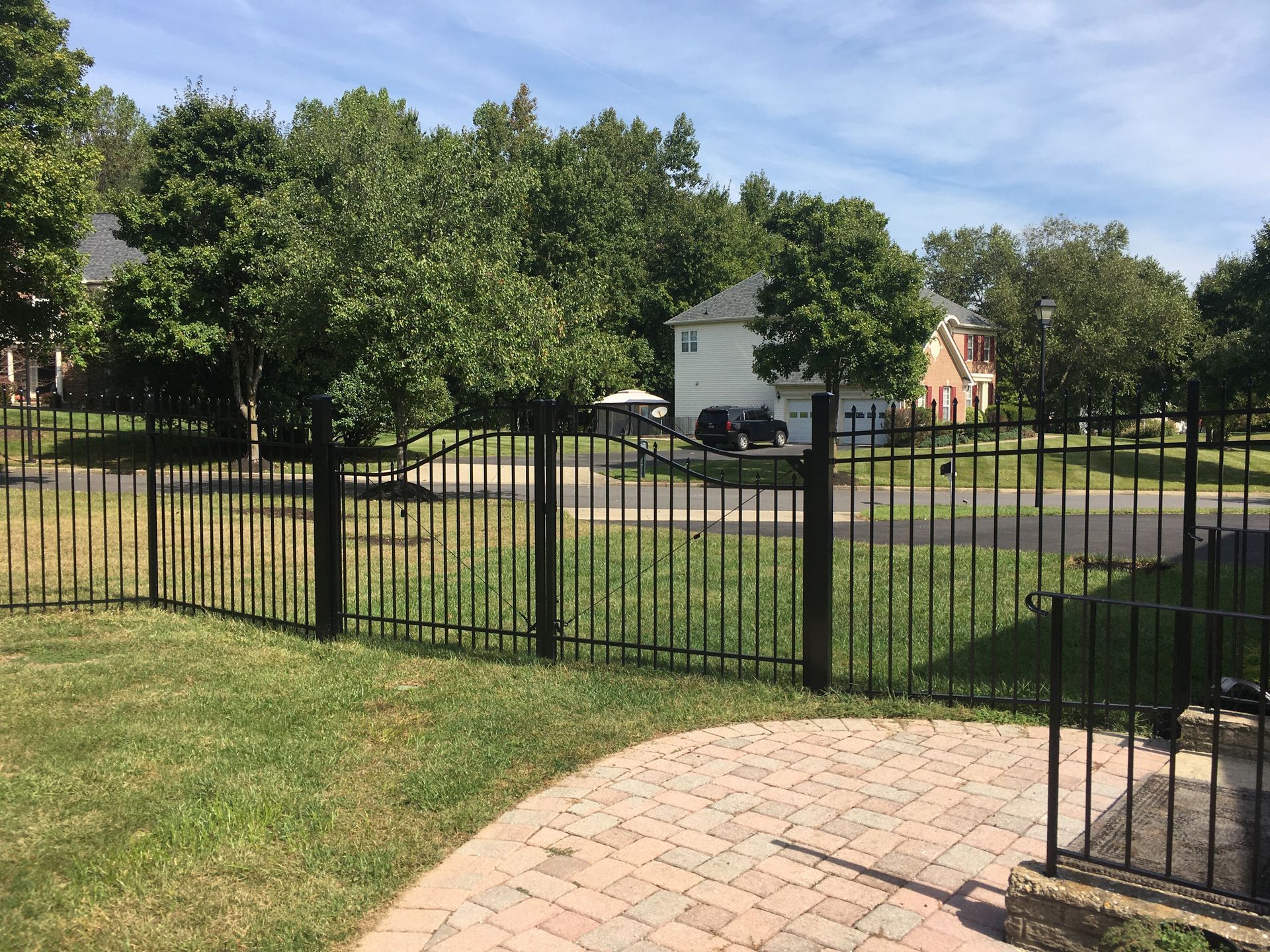 A black metal fence encloses a backyard with a brick patio and grass, with a residential neighborhood in the background.